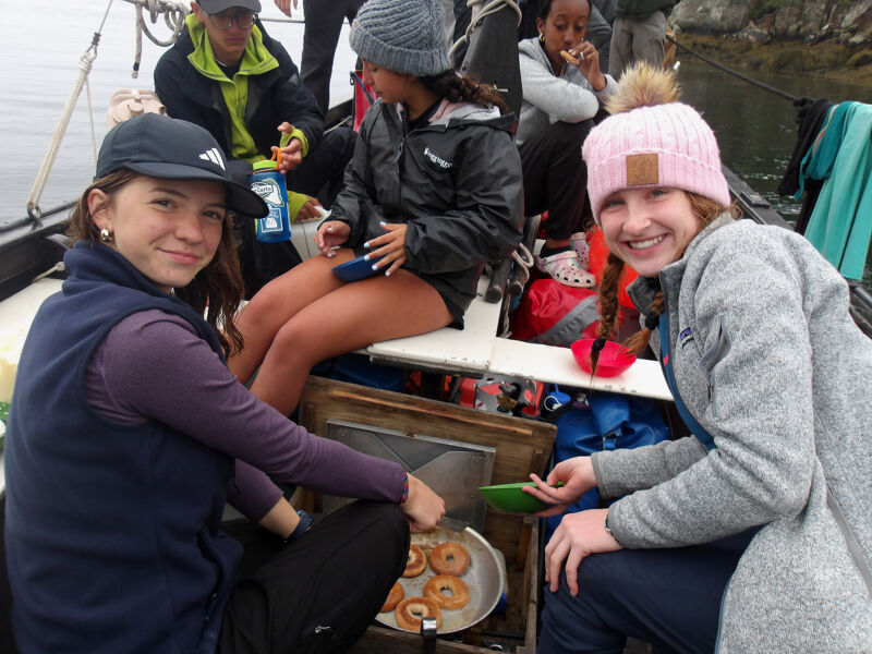 The image shows a group of young people on a boat, possibly enjoying a meal or snack. In the foreground, two girls are seated, one holding a green bowl and the other near a cooking device with what appears to be donuts. Other individuals are visible in the background, some eating. The setting suggests an outdoor adventure or excursion on the water.